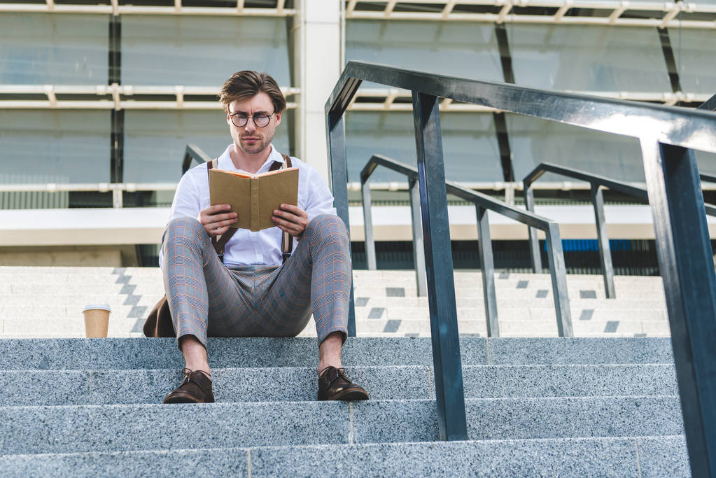 κάτω όψη του όμορφος νεαρός άνδρας με το χαρτί κύπελλο καφέ ανάγνωση του βιβλίου στις σκάλες σε δρόμο της πόλης - Φωτογραφία, εικόνα