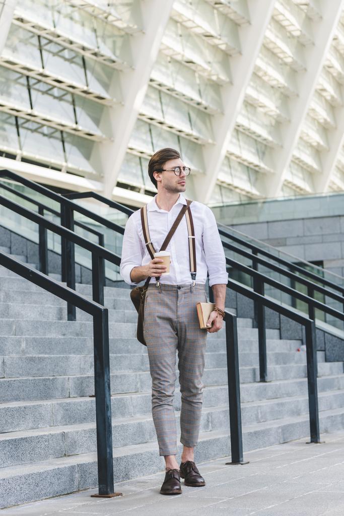 attractive young man walking in front of stadium with book and coffee to go - Photo, Image