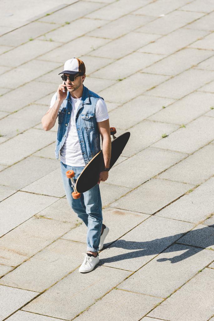 hoge hoekmening van knappe jonge skateboarder wandelen met longboard in de hand en praten via de telefoon - Foto, afbeelding