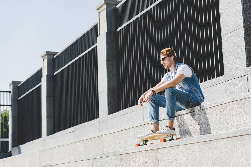 stijlvolle jongeman in denim kleding zitten op trappen met longboard weg op zoek - Foto, afbeelding