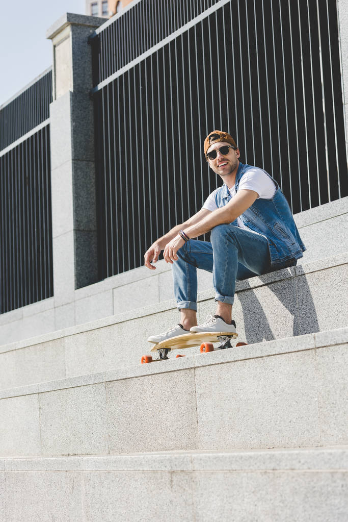 Onderaanzicht van de knappe jonge schaatser zittend op trap met longboard - Foto, afbeelding