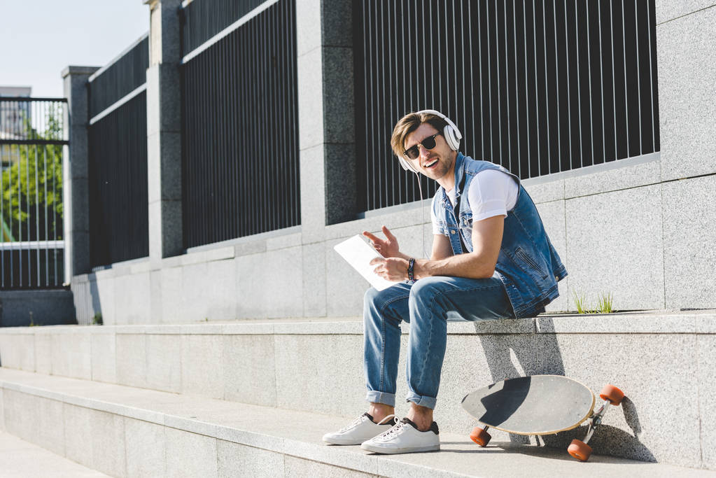 happy young man with skateboard listening music with headphones and tablet on street - Photo, Image