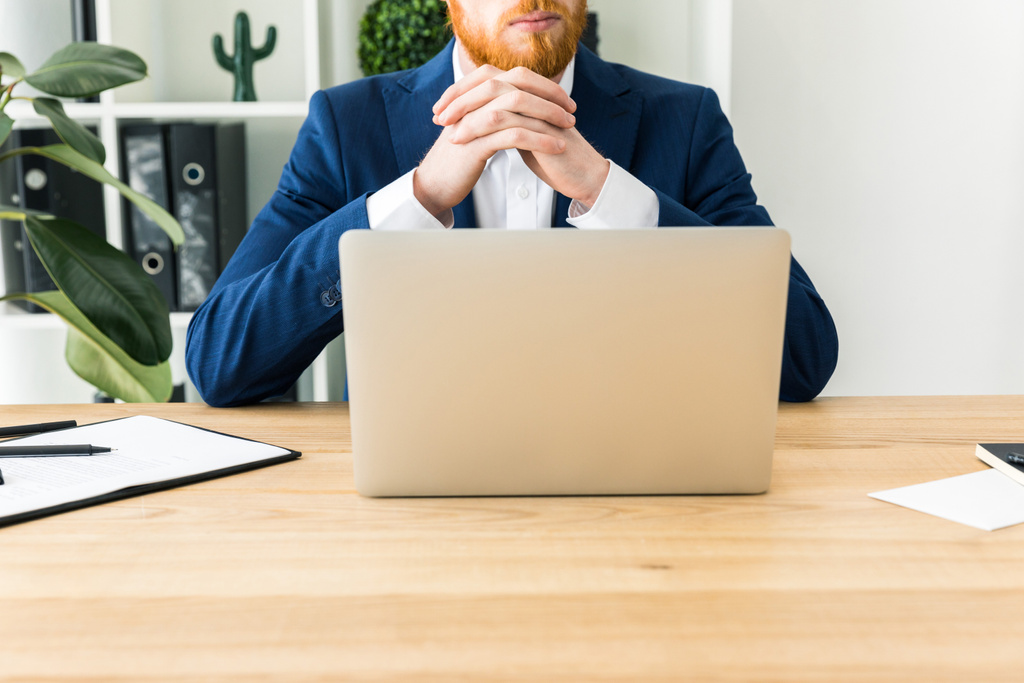 vista parcial del hombre de negocios barbudo en traje sentado en el lugar de trabajo con el ordenador portátil en la oficina
 - Foto, imagen