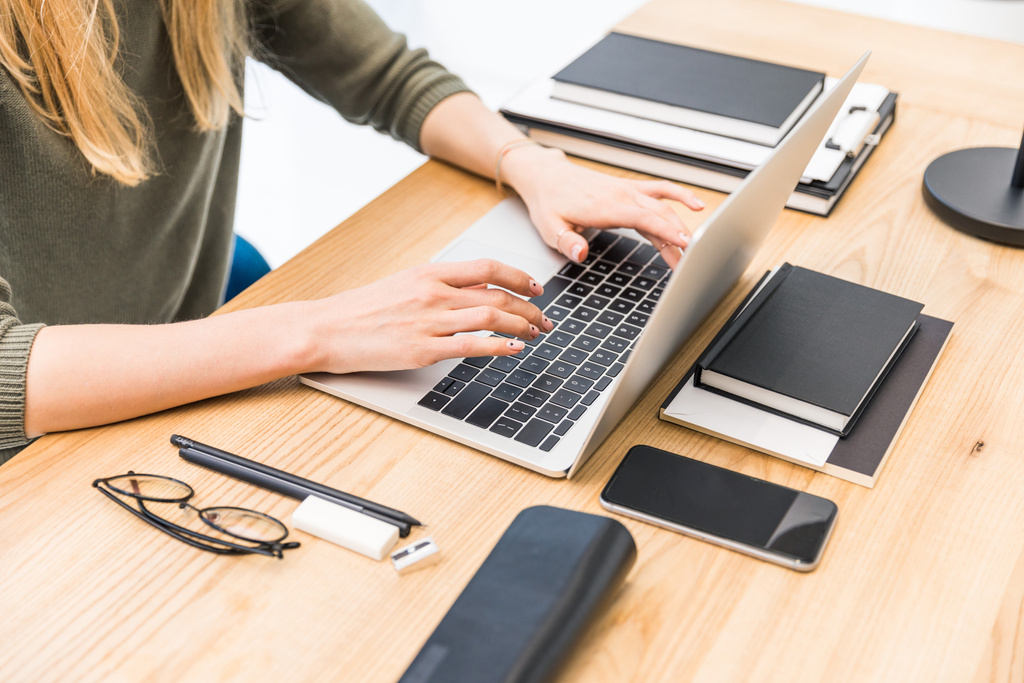 Teilbild einer Geschäftsfrau, die am Arbeitsplatz mit Notebooks, Brille und Smartphone im Büro am Laptop tippt - Foto, Bild