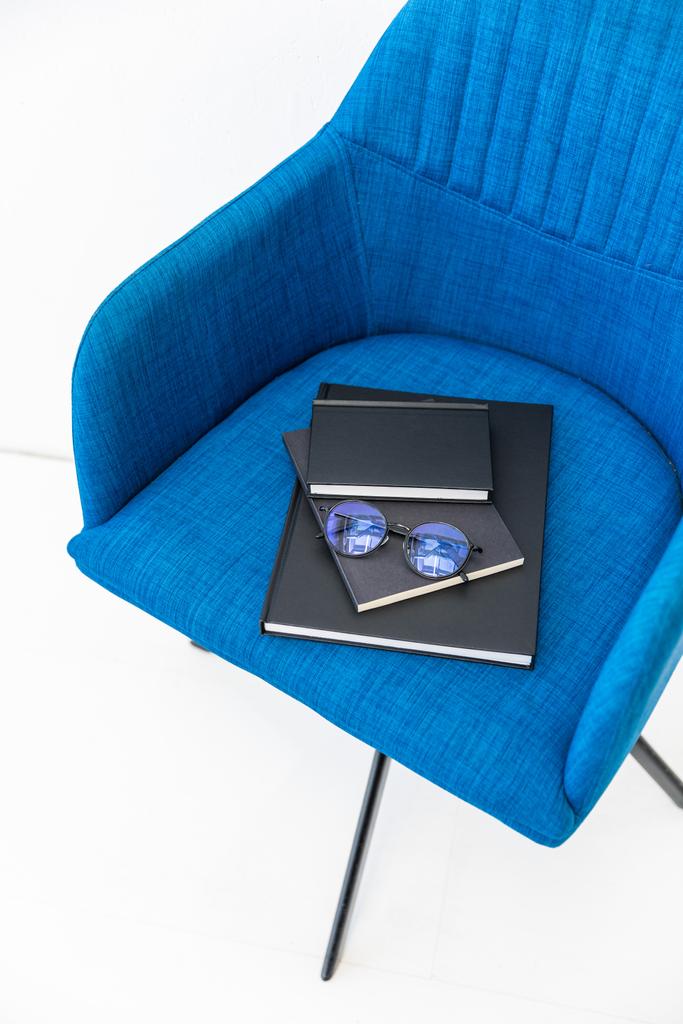 Nahaufnahme eines Stapels schwarzer Notizbücher und Brillen auf blauem Stuhl auf weißem Hintergrund - Foto, Bild