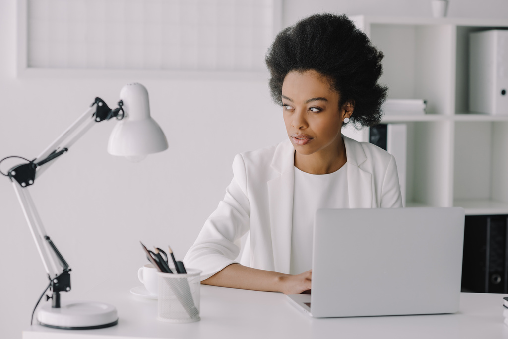 séduisante femme d'affaires afro-américaine assise à table avec ordinateur portable et regardant loin dans le bureau
 - Photo, image