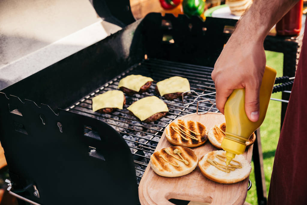 Chef adding mustard to burgers cooked outdoors on grill - Photo, Image