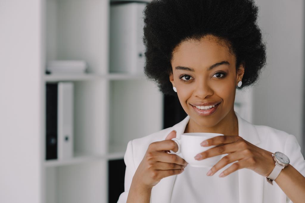 attraktive afrikanisch-amerikanische Geschäftsfrau hält eine Tasse Kaffee in der Hand und blickt in die Kamera im Büro - Foto, Bild
