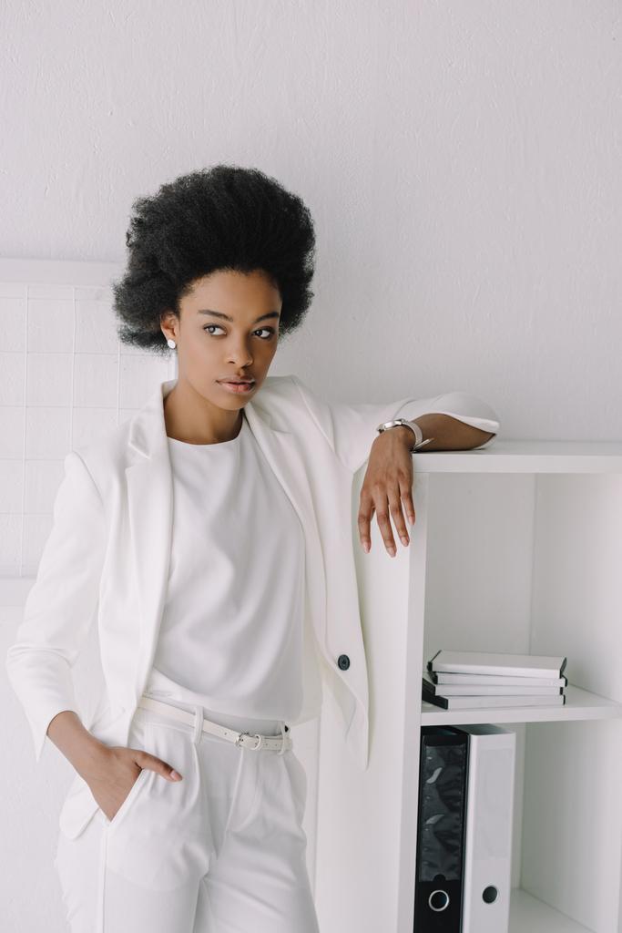 atractiva mujer de negocios afroamericana apoyada en estantes y mirando hacia otro lado en la oficina
 - Foto, imagen