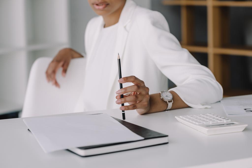 imagem recortada de mulher de negócios afro-americana sentada à mesa e segurando lápis no escritório
 - Foto, Imagem