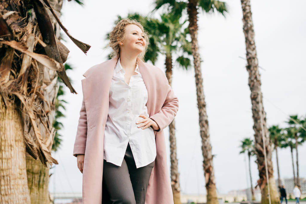 göndör fiatal nő, egy rózsaszín kabát sétál át a tavaszi parkban, és élvezi a meleg időjárás, nevet - Fotó, kép
