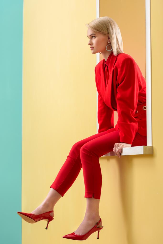 vista lateral da bela mulher loira em elegante terno vermelho e sapatos sentados na janela decorativa
 - Foto, Imagem