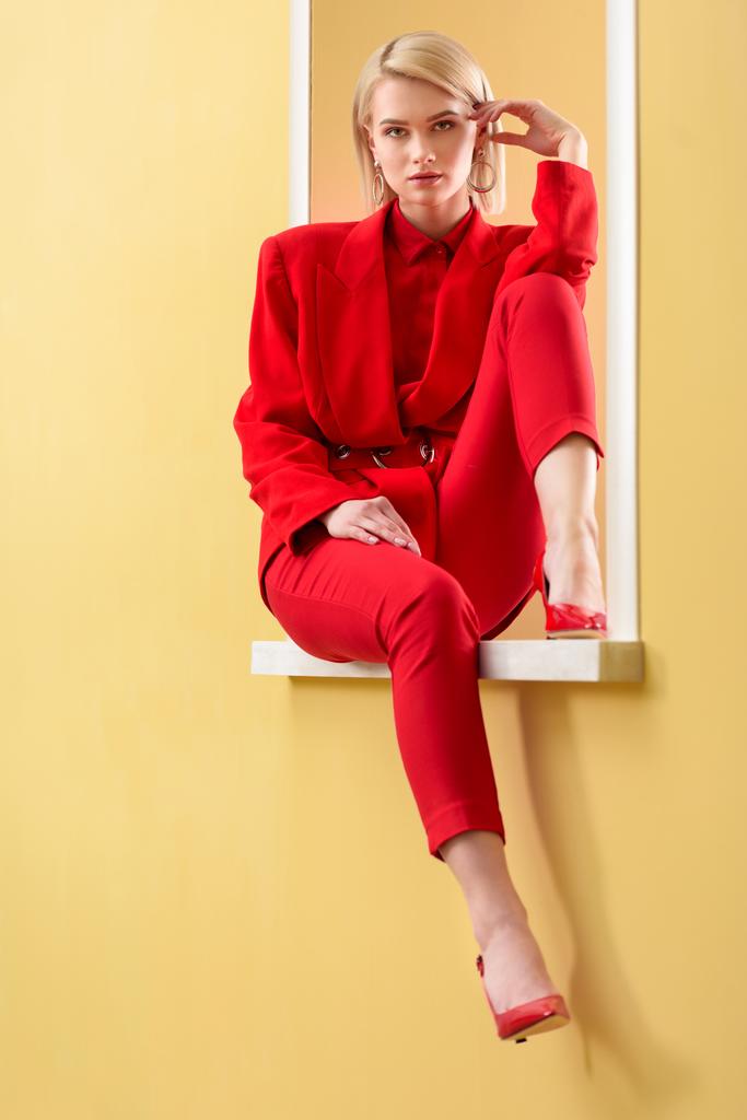красивая блондинка в стильном красном костюме и туфлях сидит на декоративном окне
 - Фото, изображение