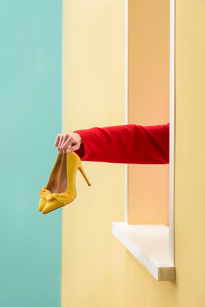 Μερική άποψη της γυναίκας που κρατά κίτρινα παπούτσια στο χέρι στον πολύχρωμο φόντο - Φωτογραφία, εικόνα