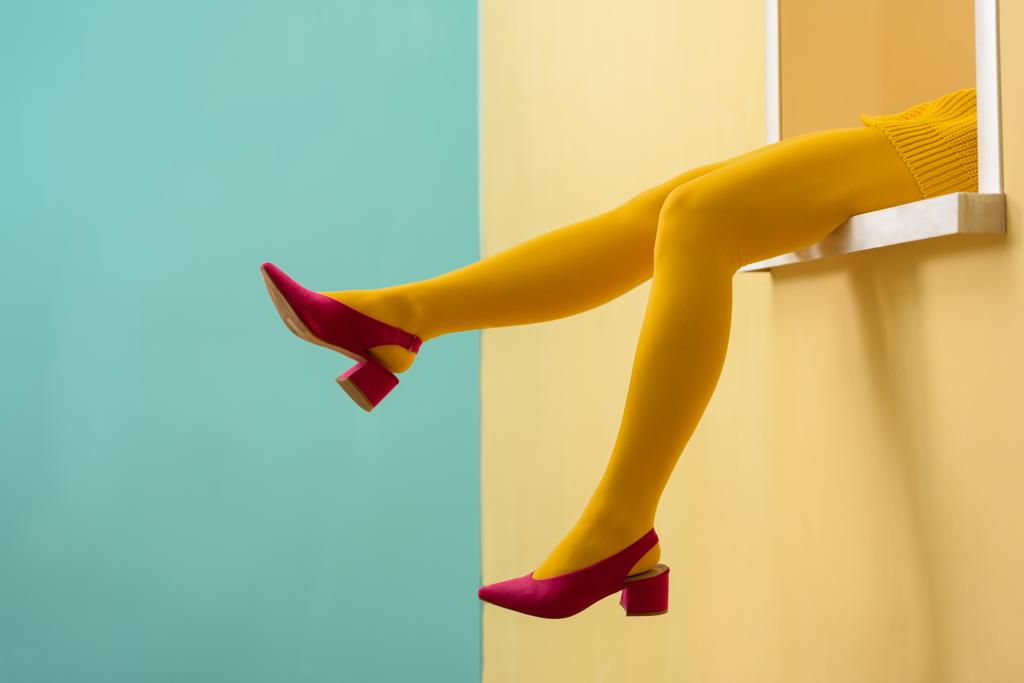 частичный вид женщины в желтых колготках и розовых туфлях, сидящей на декоративном окне на синем фоне
  - Фото, изображение