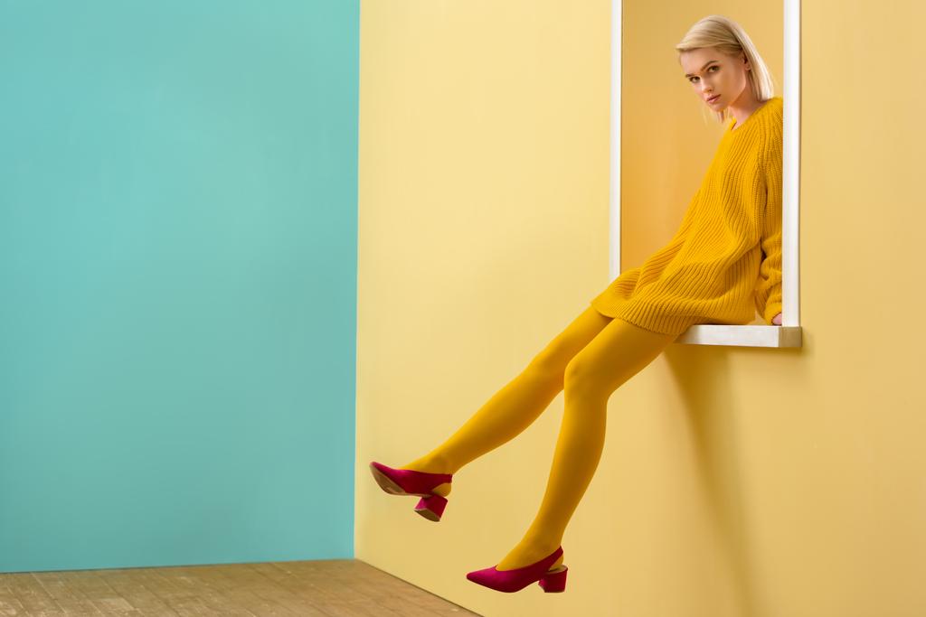 вид сбоку стильной женщины в розовых туфлях, желтом свитере и колготках, сидящей на декоративном витрине
 - Фото, изображение