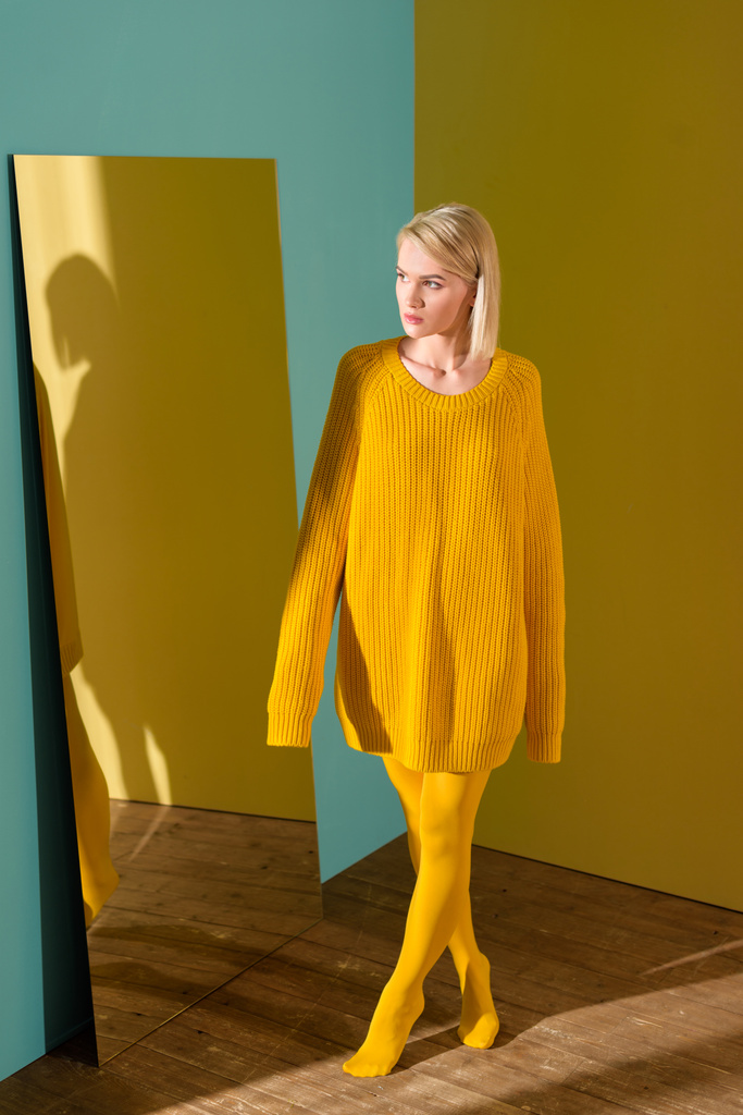 bela mulher loira pensativa em camisola amarela e meia-calça em pé no espelho
 - Foto, Imagem