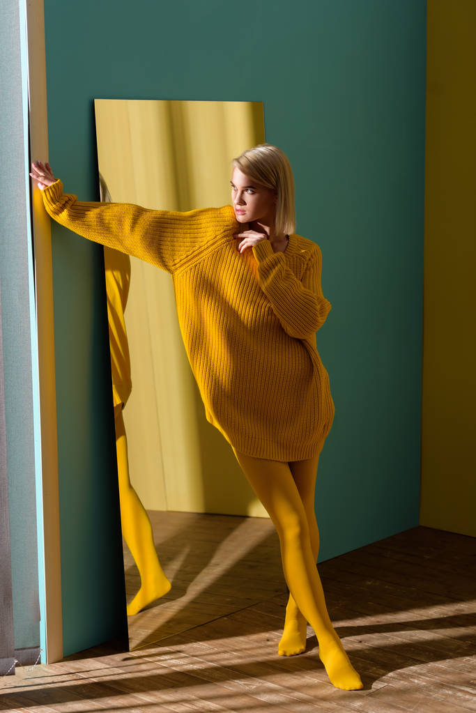 黄色のセーターとミラーに立っているタイツのスタイリッシュなブロンド女性 - 写真・画像