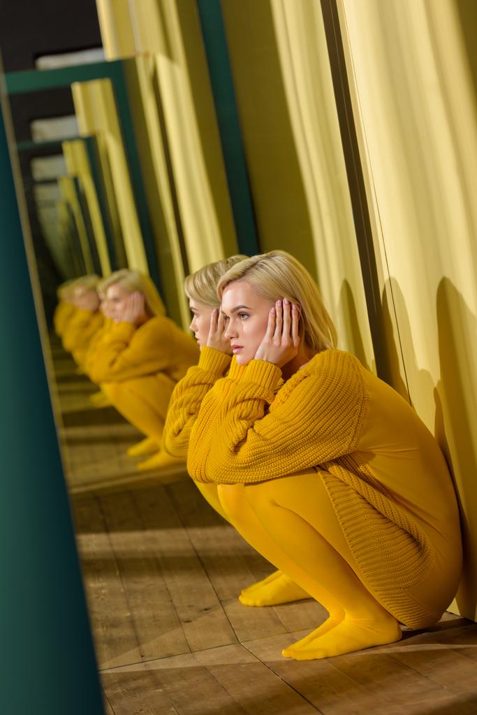 Seitenansicht der schönen, nachdenklichen Frau im gelben Pullover, die am Spiegel sitzt und ihr Spiegelbild darin hat - Foto, Bild