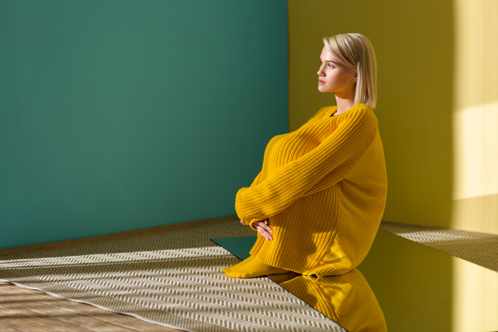 vista laterale della bella donna pensierosa in maglione giallo e collant seduti sullo specchio con riflesso in esso
 - Foto, immagini