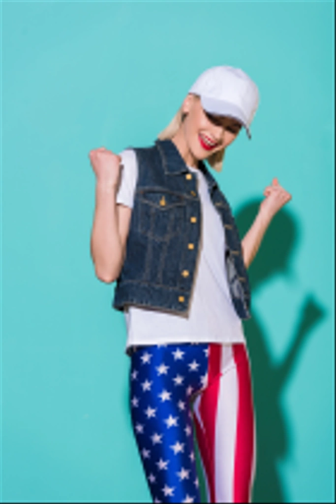 mujer alegre y elegante en gorra, camisa blanca, chaqueta de mezclilla y leggings con patrón de bandera americana posando sobre fondo azul
 - Foto, Imagen