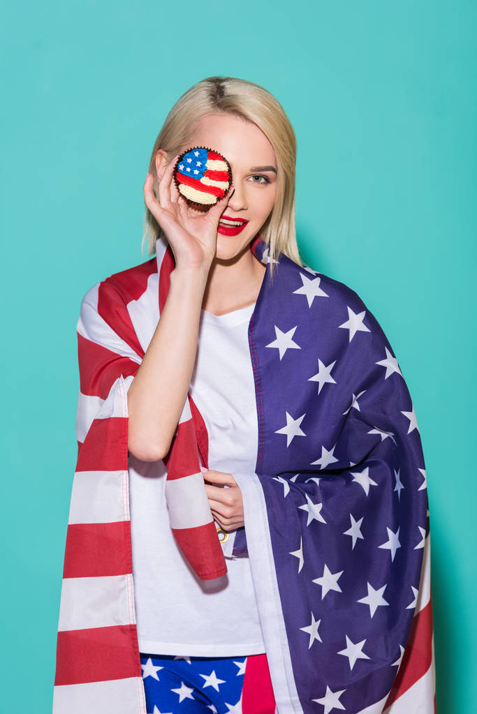 Porträt einer jungen Frau mit amerikanischer Flagge und Cupcake auf blauem Hintergrund, die den 4. Juli feiert - Foto, Bild