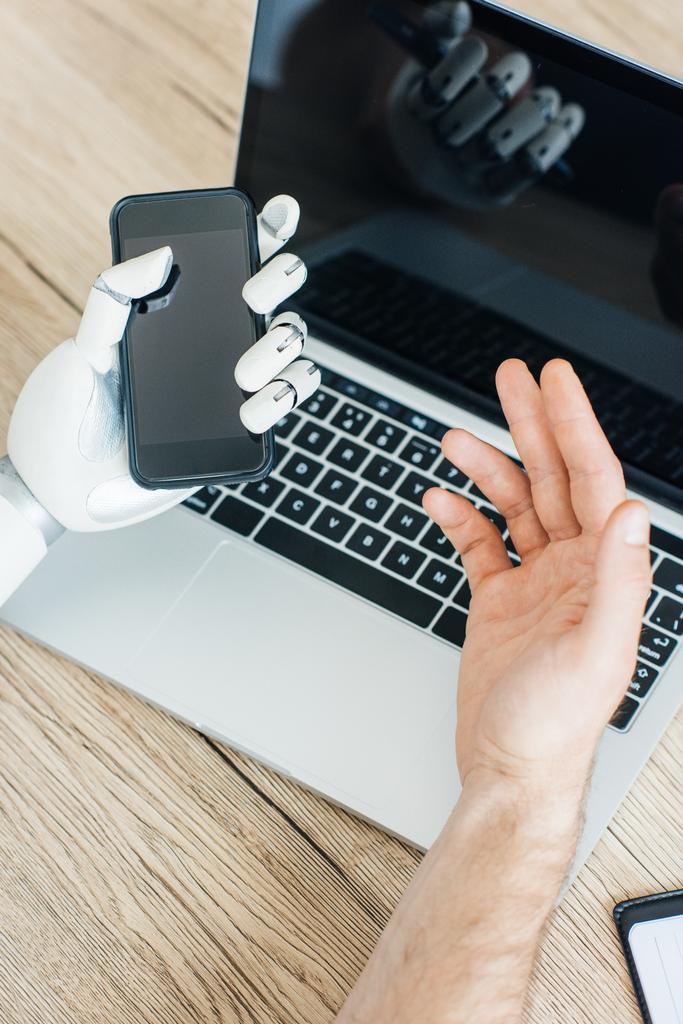 крупним планом вигляд рук людини і робота, використовуючи смартфон і ноутбук за дерев'яним столом
  - Фото, зображення