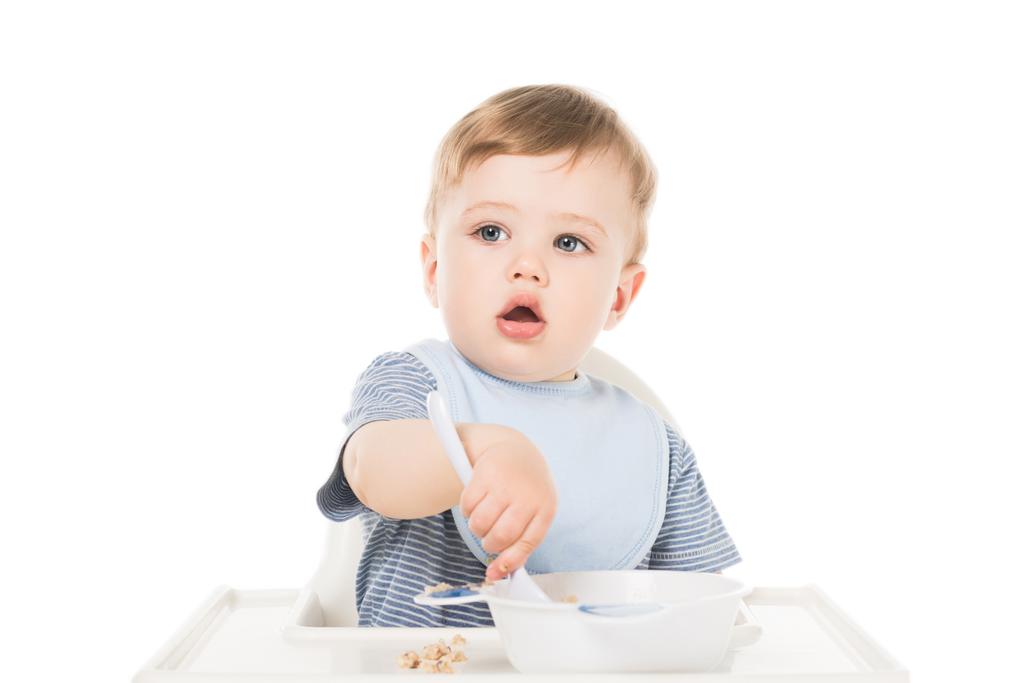 mały chłopiec w szelkach siedzi w krzesełko do karmienia i jedzenia przez łyżka na białym tle  - Zdjęcie, obraz