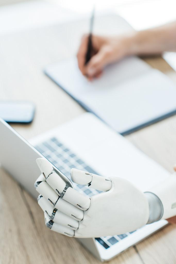 Крупный план роботизированной руки с помощью ноутбука и человеческой руки, делающей заметки на рабочем месте
 - Фото, изображение