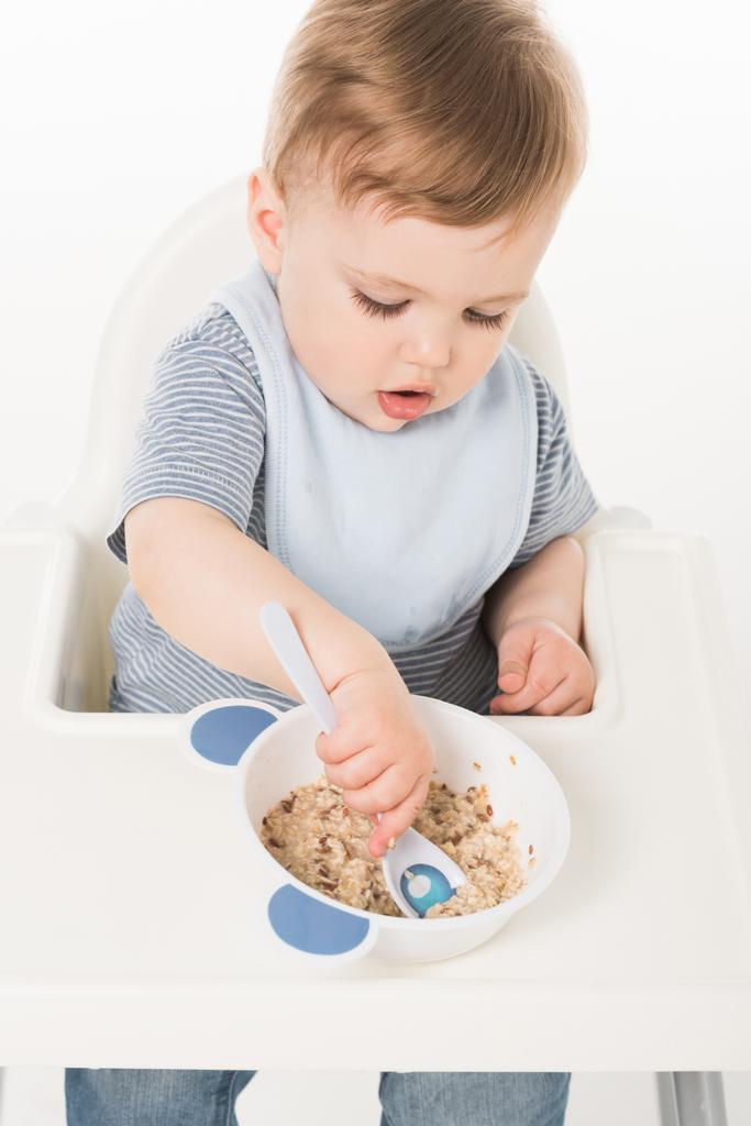 vista de ángulo alto del bebé niño en babero comiendo y sentado silla alta aislado sobre fondo blanco
  - Foto, imagen
