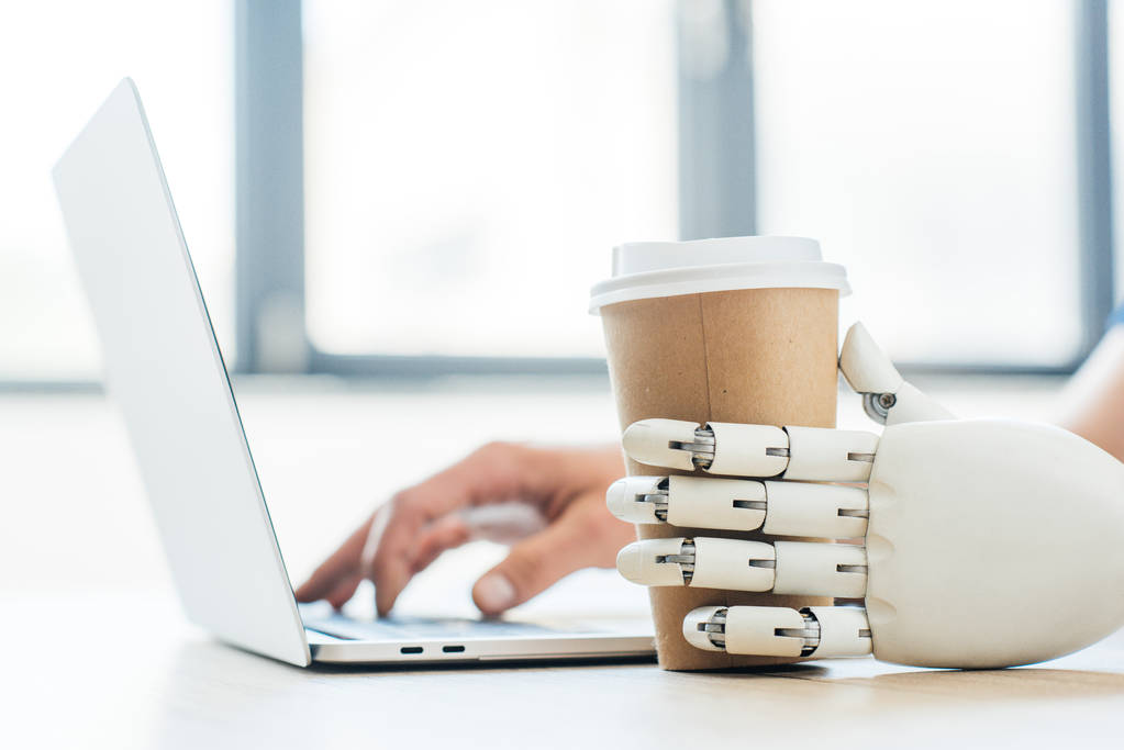 使い捨てのコーヒー カップとノート パソコンを使用して人間の手を握ったロボット アームのクローズ アップ ビュー   - 写真・画像
