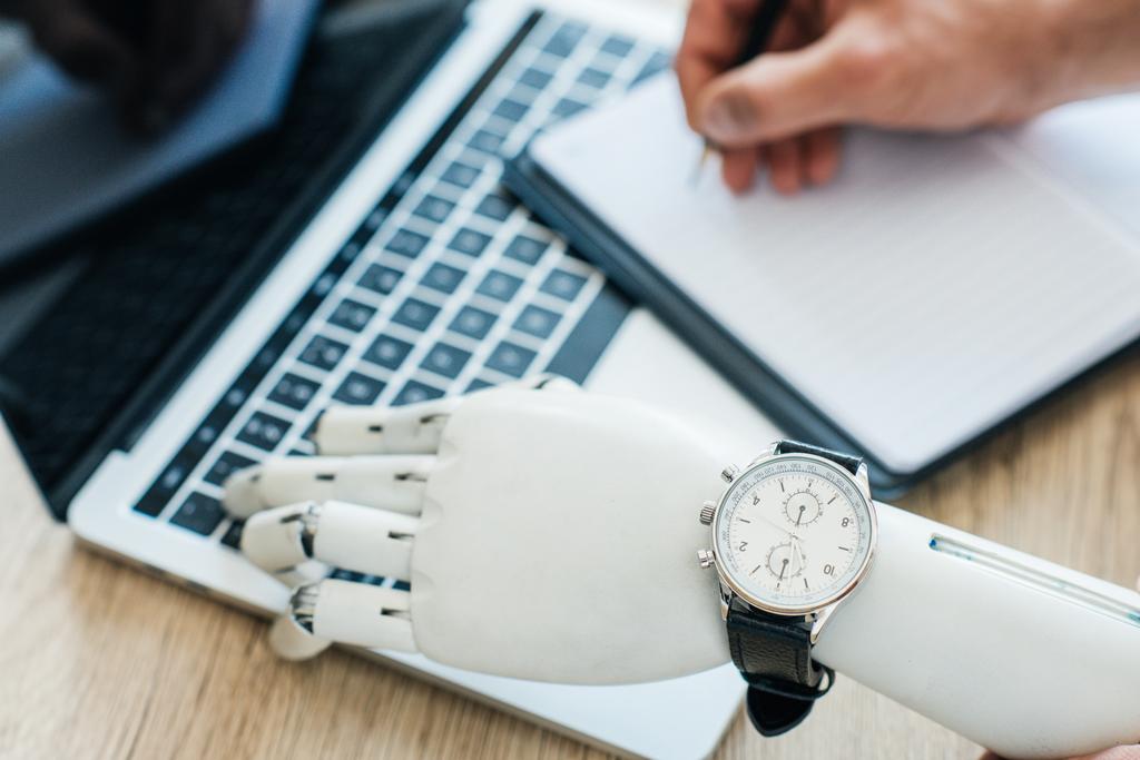 selective focus of robotic arm with wristwatch using laptop and human hand taking notes at wooden table   - Photo, Image