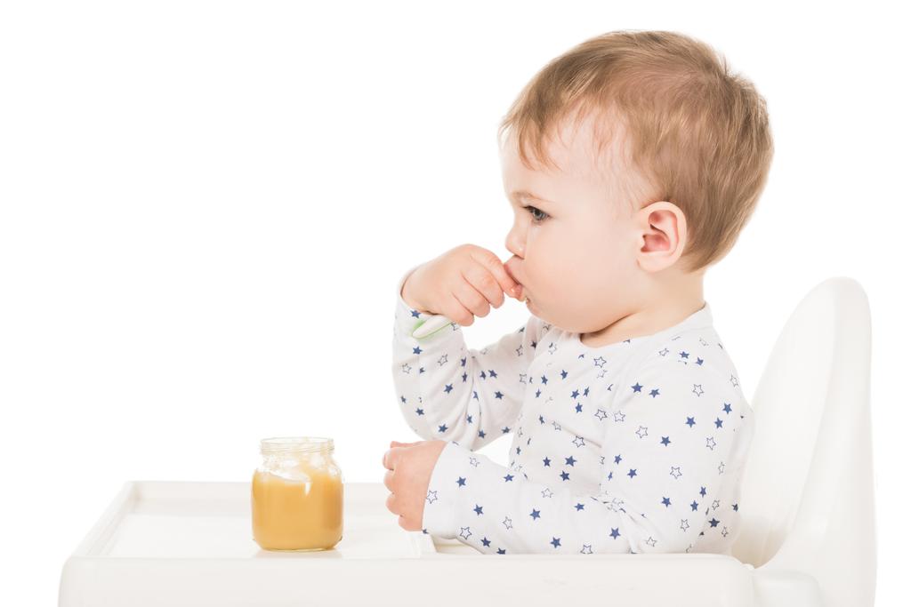  oldalnézetből a baba fiú püré jar-ból eszik, és ül, a magasszék elszigetelt fehér background  - Fotó, kép