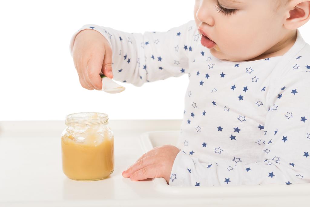  babyjongen eten puree uit de pot en zit in kinderstoel geïsoleerd op witte achtergrond  - Foto, afbeelding