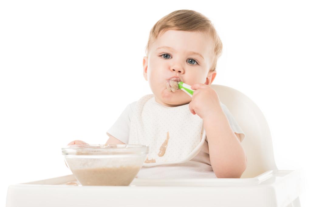 petit garçon en bavoir manger du porridge par cuillère et assis dans une chaise haute isolé sur fond blanc
  - Photo, image