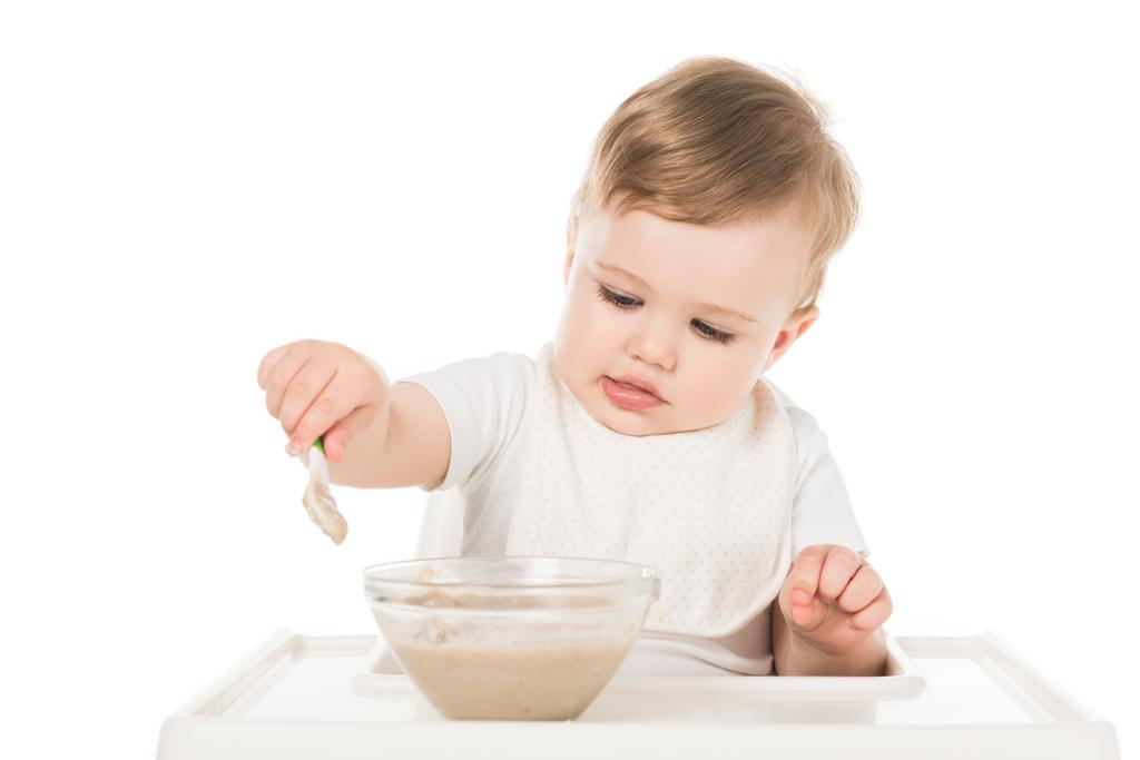 pieni poika ruokalapussa syö vauvanruokaa lusikalla ja istuu syöttötuolissa eristettynä valkoisella taustalla
  - Valokuva, kuva