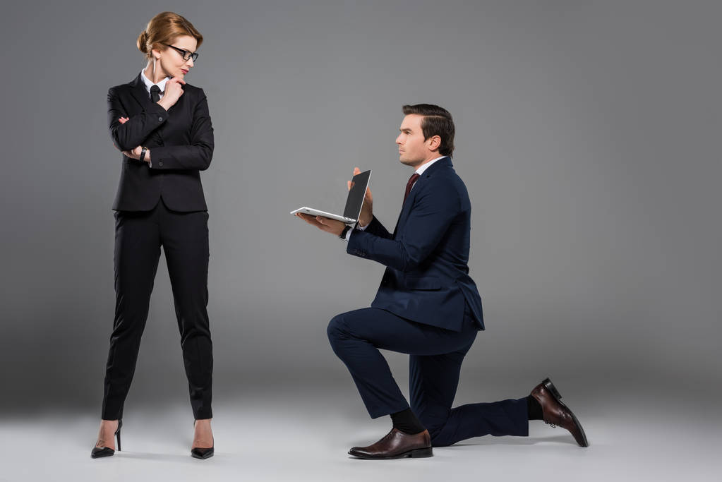 бізнесмен на одному коліні тримає ноутбук для феміністської бізнес-леді, ізольований на сірому
 - Фото, зображення
