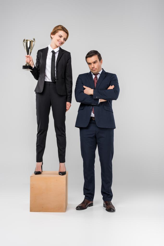 feliz mujer de negocios con copa de trofeo de pie en el podio, molesto hombre de negocios de pie cerca, aislado en gris
 - Foto, Imagen