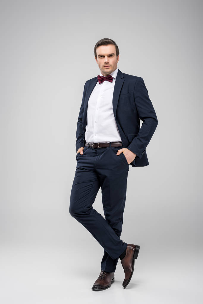 handsome man posing in tuxedo with hands in pockets, isolated on grey - Photo, Image