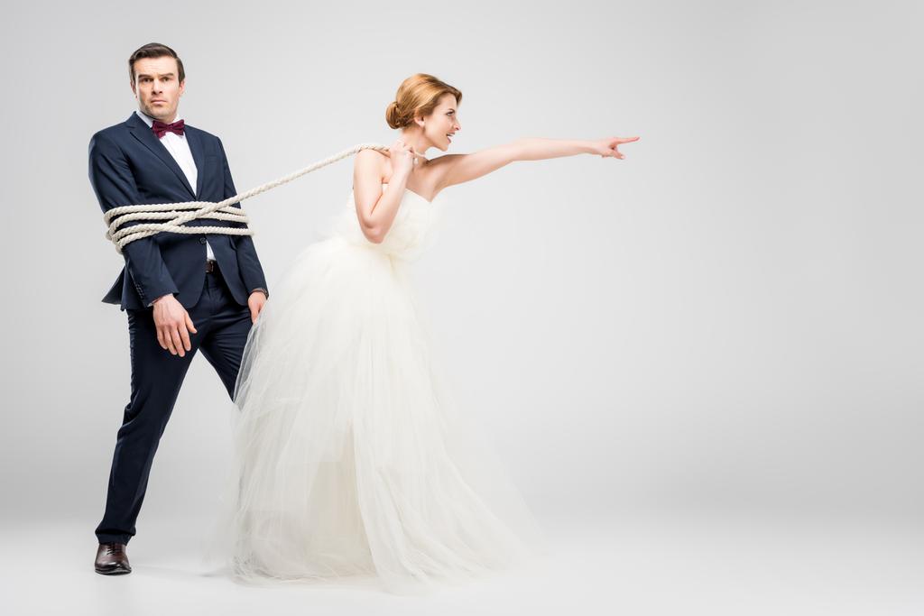 Braut zeigt und zieht Bräutigam mit Seil gebunden, isoliert auf grau, Feminismus-Konzept - Foto, Bild
