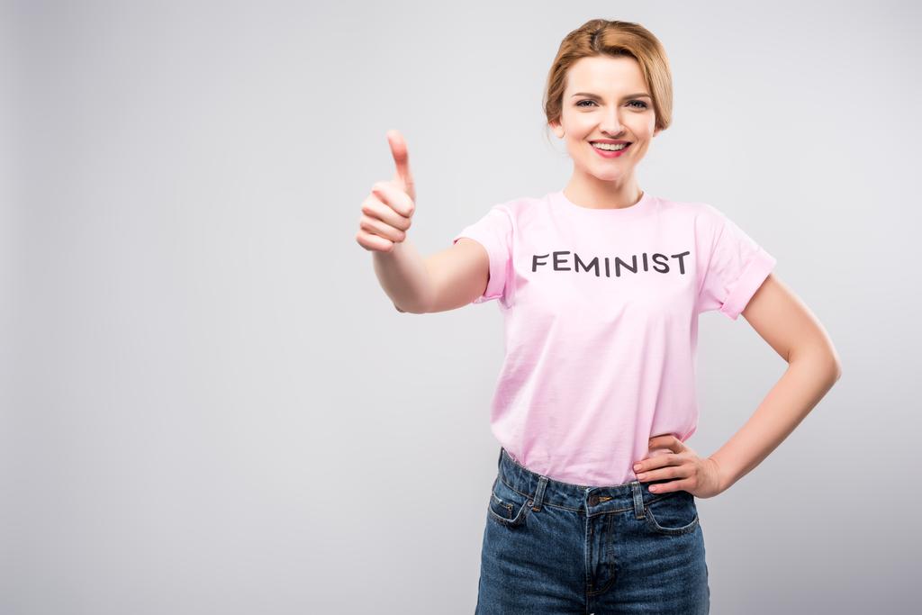 uśmiechający się kobieta w różowy feministycznej t-shirt wyświetlone kciuk w górę, na szarym tle - Zdjęcie, obraz