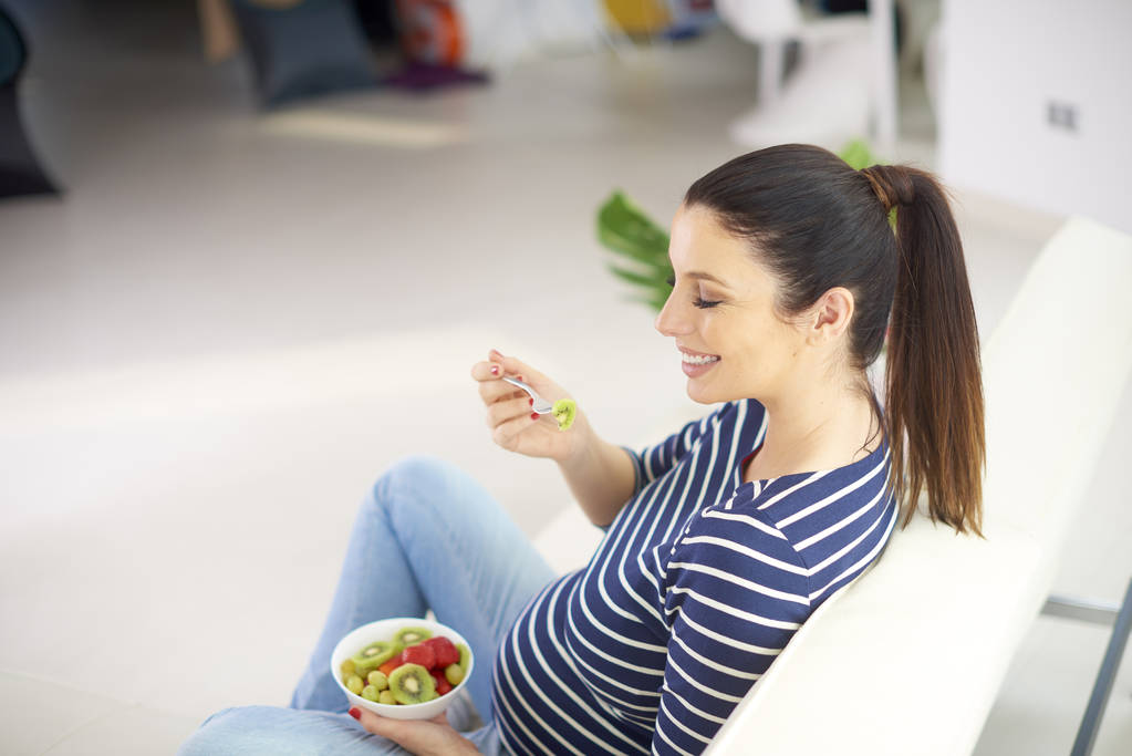 Високо під кутом знімок красивої вагітної жінки, що розслабляється вдома і їсть миску фруктового салату
. - Фото, зображення