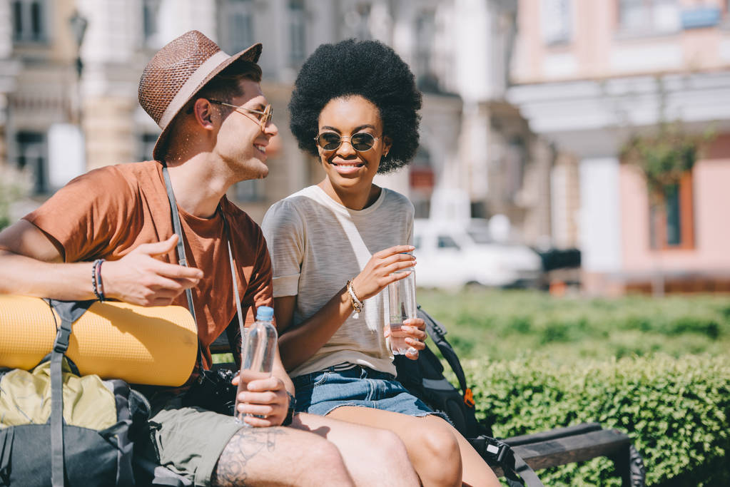 happy interracial couple of tourists with bottles of water sitting on bench  - Photo, Image