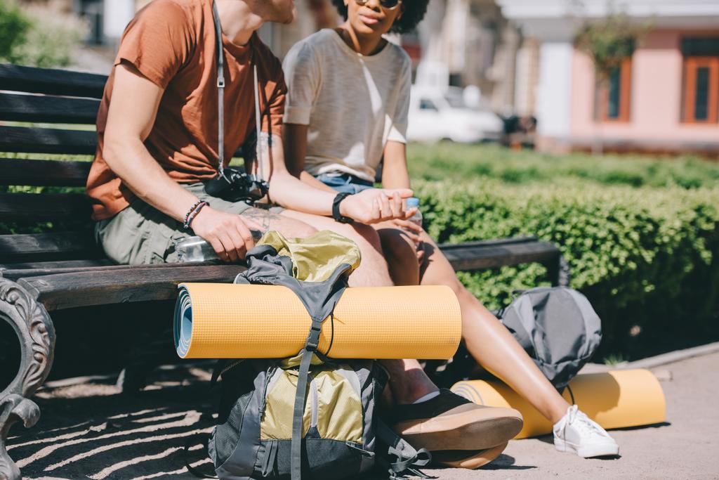 imagem cortada de casal de viajantes sentados no banco com mochilas e tapetes
  - Foto, Imagem