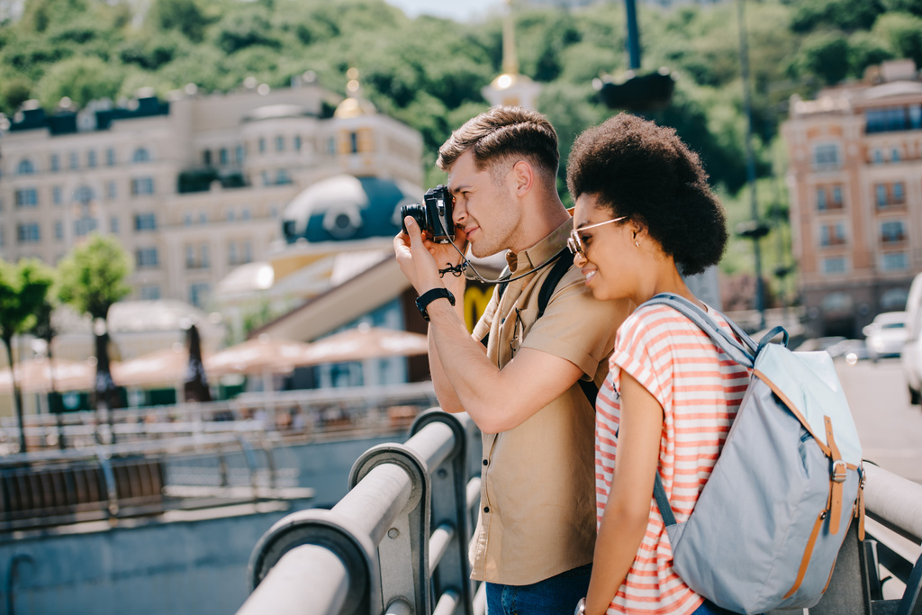 widok boczny podróżnik mężczyzna biorąc obraz na aparat i african american dziewczyna stoi w pobliżu  - Zdjęcie, obraz
