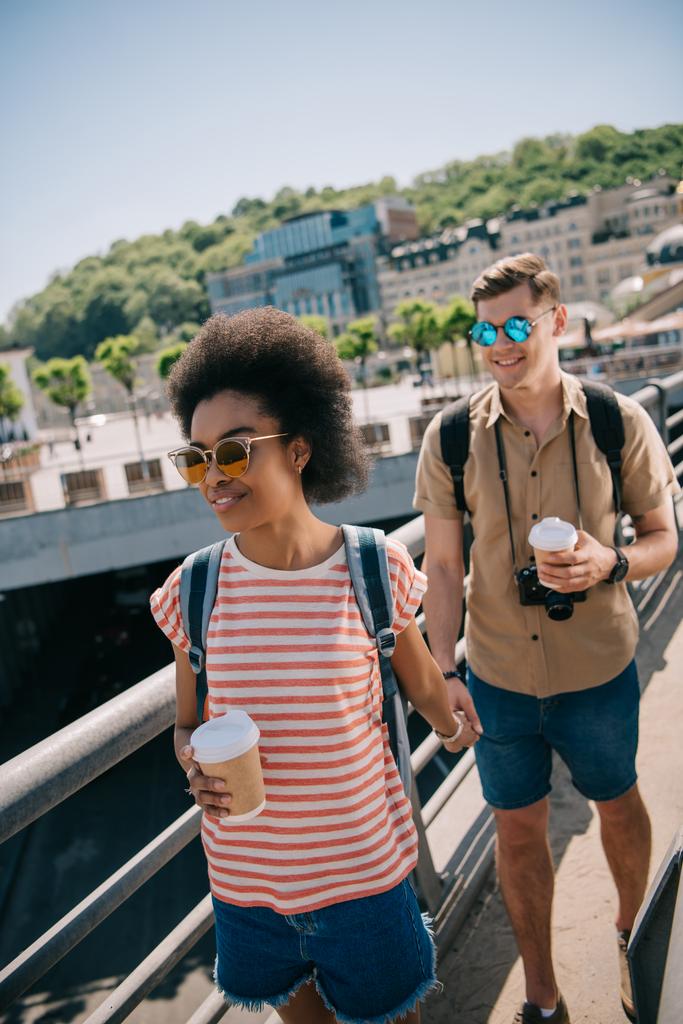 junges multikulturelles Touristenpaar mit Kaffeetassen und Kamera läuft über Brücke  - Foto, Bild