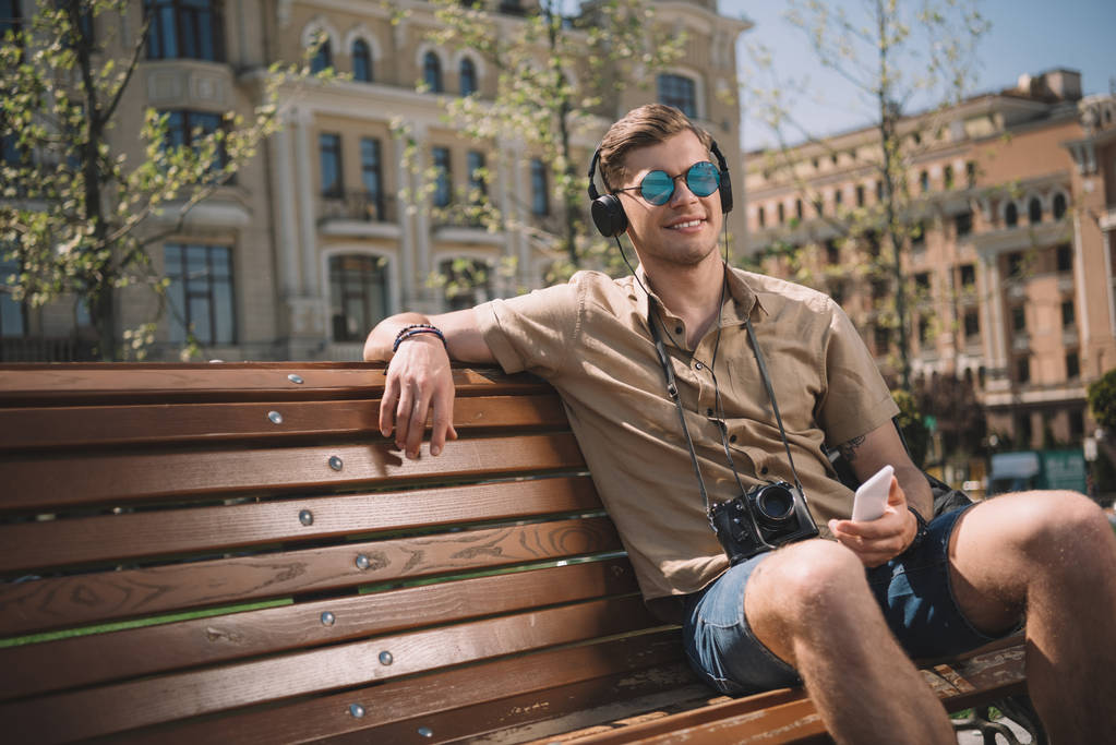 Junger Mann mit Sonnenbrille, Kamera, Kopfhörer und Smartphone sitzt auf Bank  - Foto, Bild