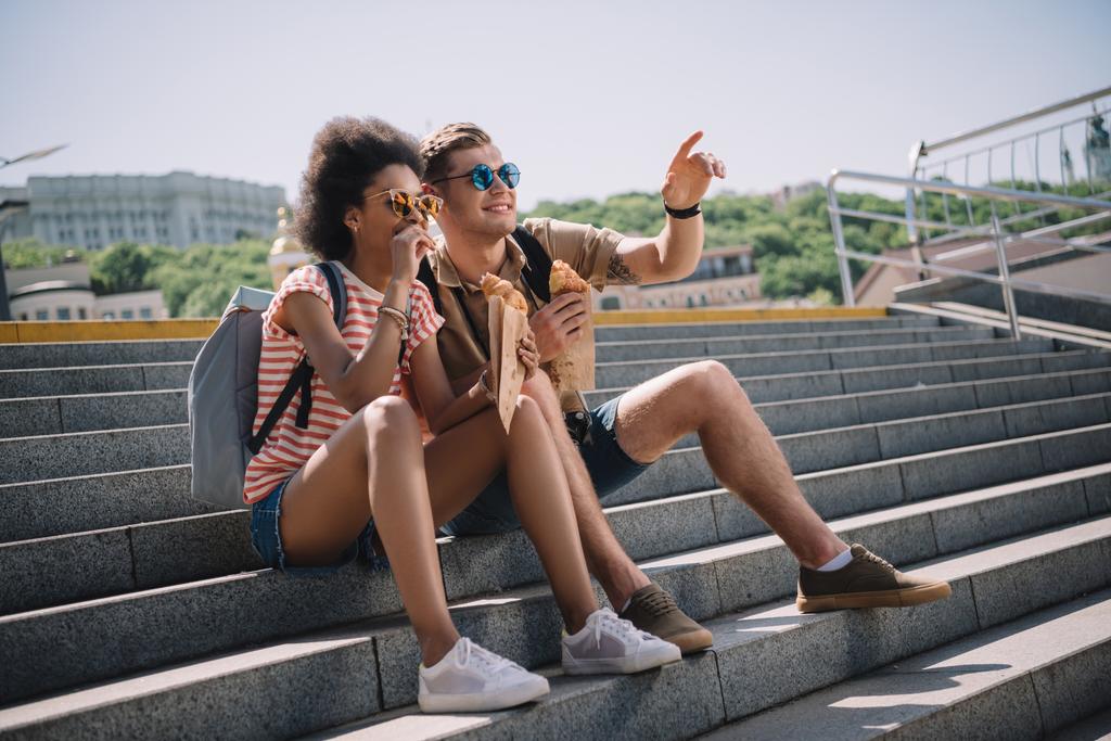 young stylish couple in sunglasses sitting on stairs and eating croissants  - Photo, Image