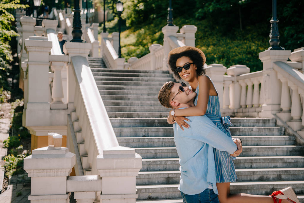 joven feliz sosteniendo novia afroamericana cerca de escaleras en el parque
  - Foto, Imagen