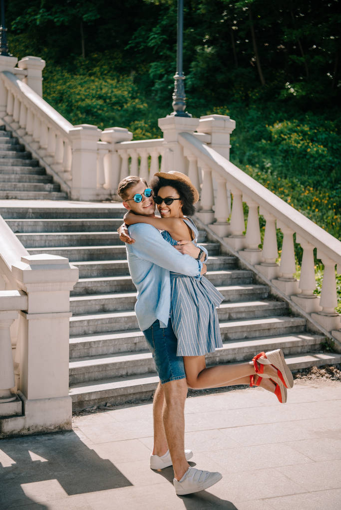 elegante hombre sosteniendo y abrazando joven afroamericano novia en parque
  - Foto, Imagen